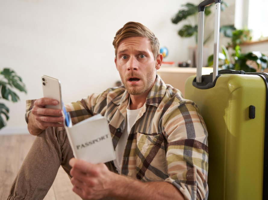 Man sitting beside luggage looking shocked while holding his phone and passport.